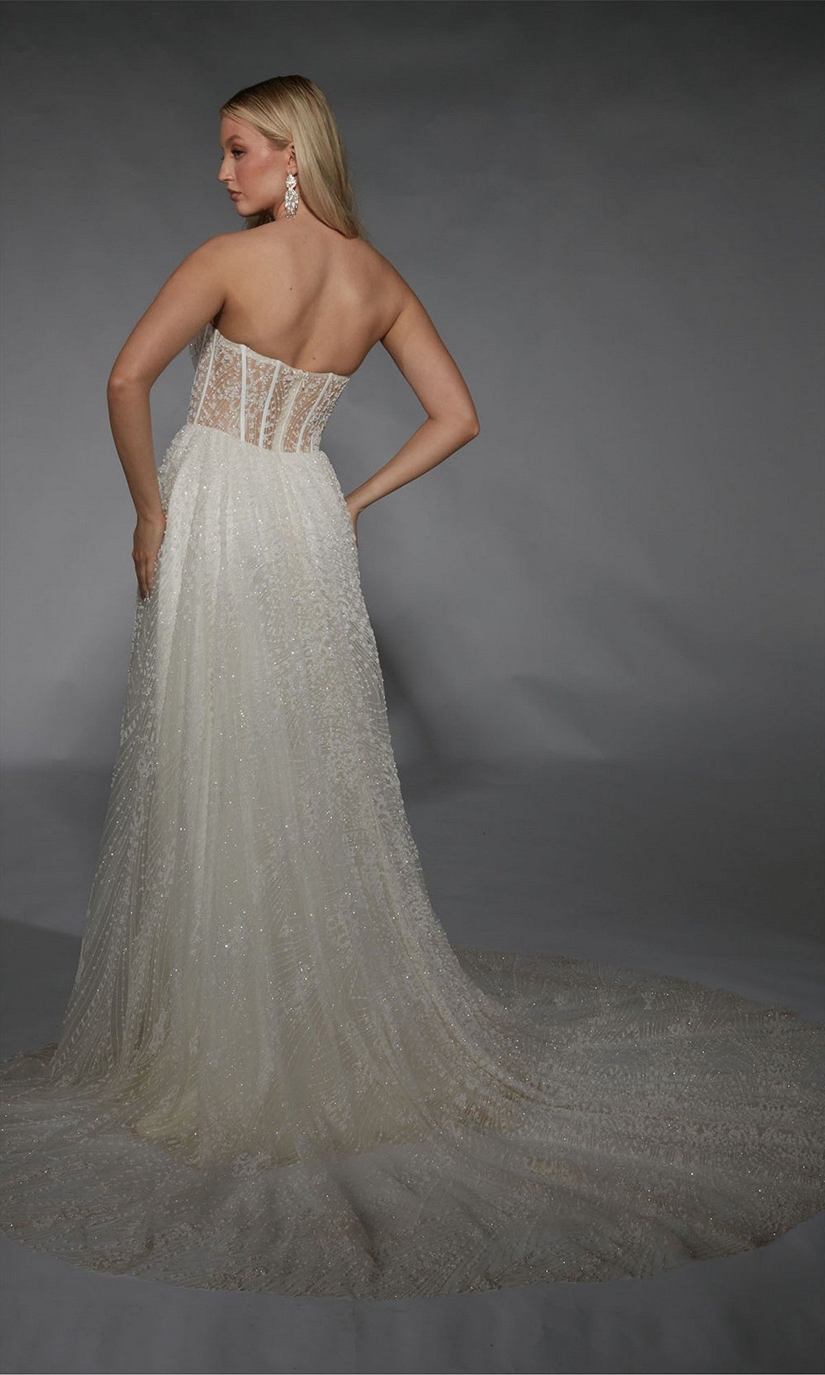 A woman poses in the Alyce 7133 long formal dress—a strapless, floor-length white gown with a sheer corset bodice and sparkling train—set against a gray background.